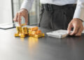 A person in business attire stacks gold coins and bars on a desk while using a calculator, suggesting financial planning or investment in gold. | MONEY6X