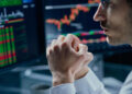 A person in a white shirt sits in front of computer monitors displaying colorful stock charts and financial data, appearing focused and contemplative with hands clasped near their face. | MONEY6X
