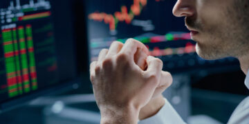 A person in a white shirt sits in front of computer monitors displaying colorful stock charts and financial data, appearing focused and contemplative with hands clasped near their face. | MONEY6X