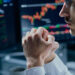 A person in a white shirt sits in front of computer monitors displaying colorful stock charts and financial data, appearing focused and contemplative with hands clasped near their face. | MONEY6X