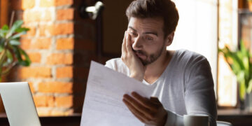 A man sits at a table with a laptop and coffee cup, holding a piece of paper and looking concerned or stressed, resting his face on his hand in a brightly lit room with brick walls and plants. | MONEY6X
