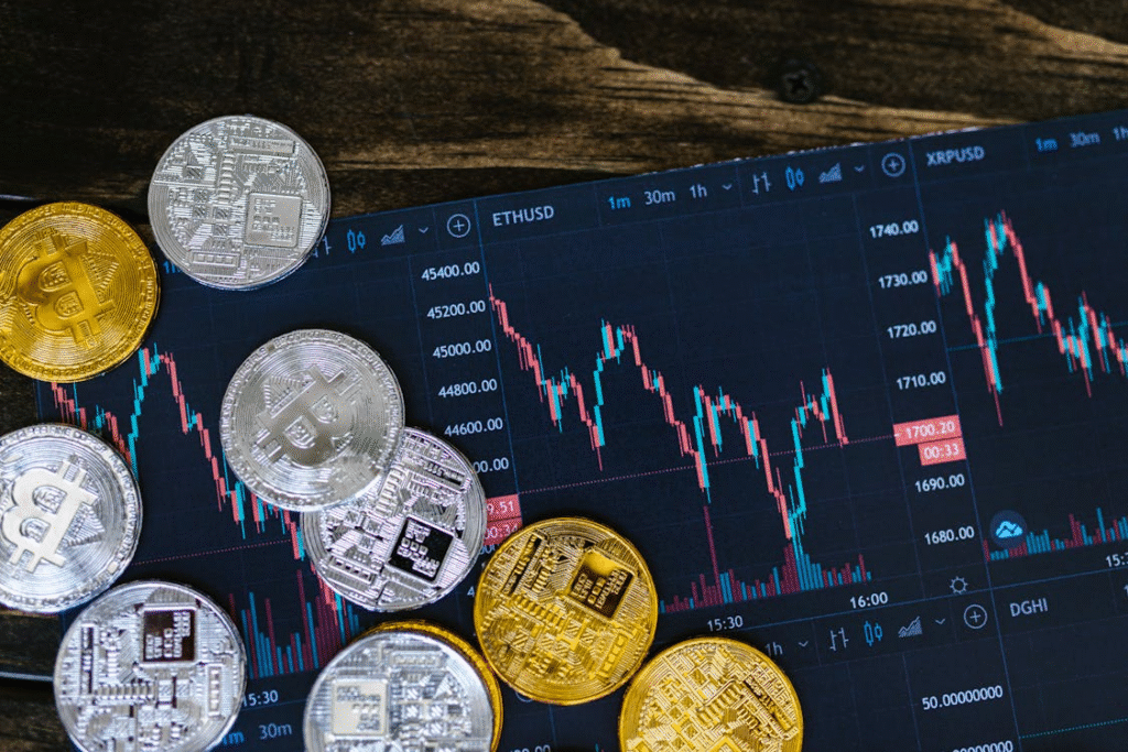 Cryptocurrency coins, including Bitcoin and Ethereum, are scattered on a surface with a digital screen in the background displaying candlestick charts and price data for various cryptocurrencies. | MONEY6X