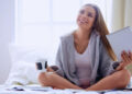 A woman sits cross-legged on a bed, smiling and looking up. She is holding a tablet in one hand and a cup in the other, wearing a cozy robe over a white shirt, with sunlight streaming in through a window behind her. | MONEY6X