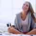 A woman sits cross-legged on a bed, smiling and looking up. She is holding a tablet in one hand and a cup in the other, wearing a cozy robe over a white shirt, with sunlight streaming in through a window behind her. | MONEY6X