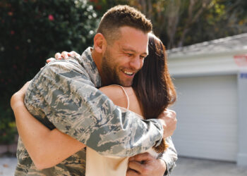 A man in military uniform smiles warmly as he hugs a woman outdoors, in front of a garage, during the daytime. | MONEY6X