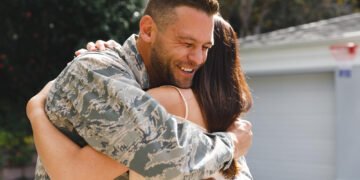 A man in military uniform smiles warmly as he hugs a woman outdoors, in front of a garage, during the daytime. | MONEY6X