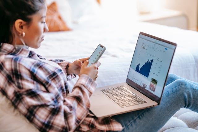 A person sits on a bed using a smartphone while a laptop on their lap displays a financial chart with a rising trend. The person wears a plaid shirt and appears focused on both devices. | MONEY6X