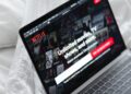 A laptop displaying the Netflix homepage with the message “Unlimited movies, TV shows, and more,” resting on a white bedspread. The Netflix logo and various show thumbnails are visible on the screen. | MONEY6X