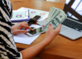 A person using a smartphone while holding several U.S. hundred-dollar bills, with a laptop, documents, and a pen on a wooden desk. | MONEY6X