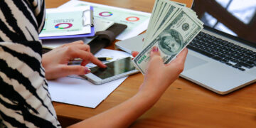A person using a smartphone while holding several U.S. hundred-dollar bills, with a laptop, documents, and a pen on a wooden desk. | MONEY6X