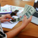 A person using a smartphone while holding several U.S. hundred-dollar bills, with a laptop, documents, and a pen on a wooden desk. | MONEY6X