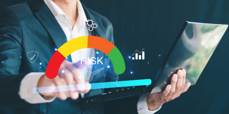 A man in a suit holds a laptop and touches a virtual risk meter, which displays a colored gauge labeled "RISK" and various digital icons, representing risk assessment or management. | MONEY6X