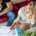 A woman and a man sit at a glass table with papers, a blue calculator, and a laptop, looking concerned as they review documents together in a living room with a red sofa. | MONEY6X