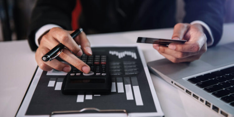 A person in business attire uses a calculator and holds a smartphone at a desk, with a clipboard displaying a bar chart and a laptop nearby, suggesting financial or business analysis. | MONEY6X