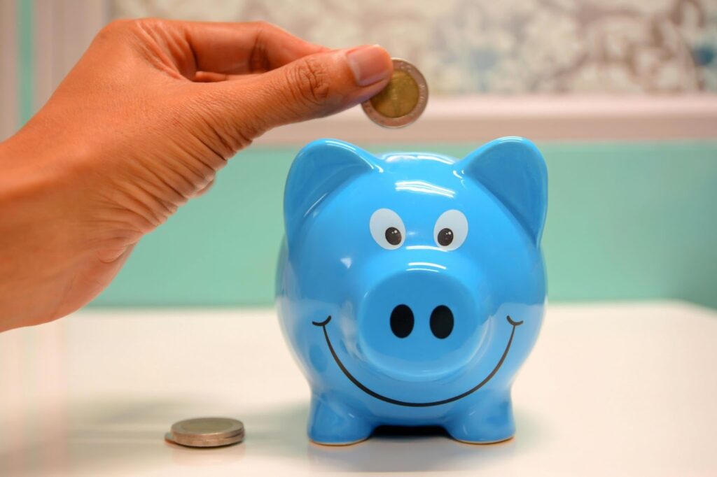 A hand holds a coin above a blue piggy bank with a smiling face; another coin rests on the white surface beside it. | MONEY6X