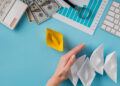 A hand blocks a row of four white paper boats, separating them from a single yellow paper boat. Nearby are a keyboard, calculator, graph paper, magnifying glass, and several $100 bills on a blue desk. | MONEY6X