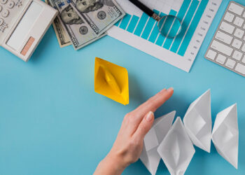 A hand blocks a row of four white paper boats, separating them from a single yellow paper boat. Nearby are a keyboard, calculator, graph paper, magnifying glass, and several $100 bills on a blue desk. | MONEY6X