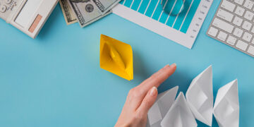 A hand blocks a row of four white paper boats, separating them from a single yellow paper boat. Nearby are a keyboard, calculator, graph paper, magnifying glass, and several $100 bills on a blue desk. | MONEY6X