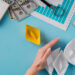 A hand blocks a row of four white paper boats, separating them from a single yellow paper boat. Nearby are a keyboard, calculator, graph paper, magnifying glass, and several $100 bills on a blue desk. | MONEY6X