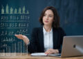 A businesswoman in a suit sits at a desk with a laptop, notebook, and books, presenting virtual financial graphs and charts with her hand, in a modern office setting. | MONEY6X