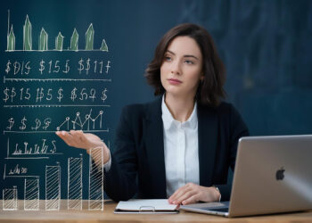 A businesswoman in a suit sits at a desk with a laptop, notebook, and books, presenting virtual financial graphs and charts with her hand, in a modern office setting. | MONEY6X