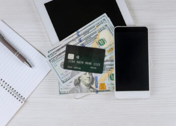 A smartphone, a tablet, a notebook with a pen, and a credit card placed on top of several hundred-dollar bills are arranged on a light wooden surface. | MONEY6X