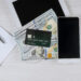 A smartphone, a tablet, a notebook with a pen, and a credit card placed on top of several hundred-dollar bills are arranged on a light wooden surface. | MONEY6X