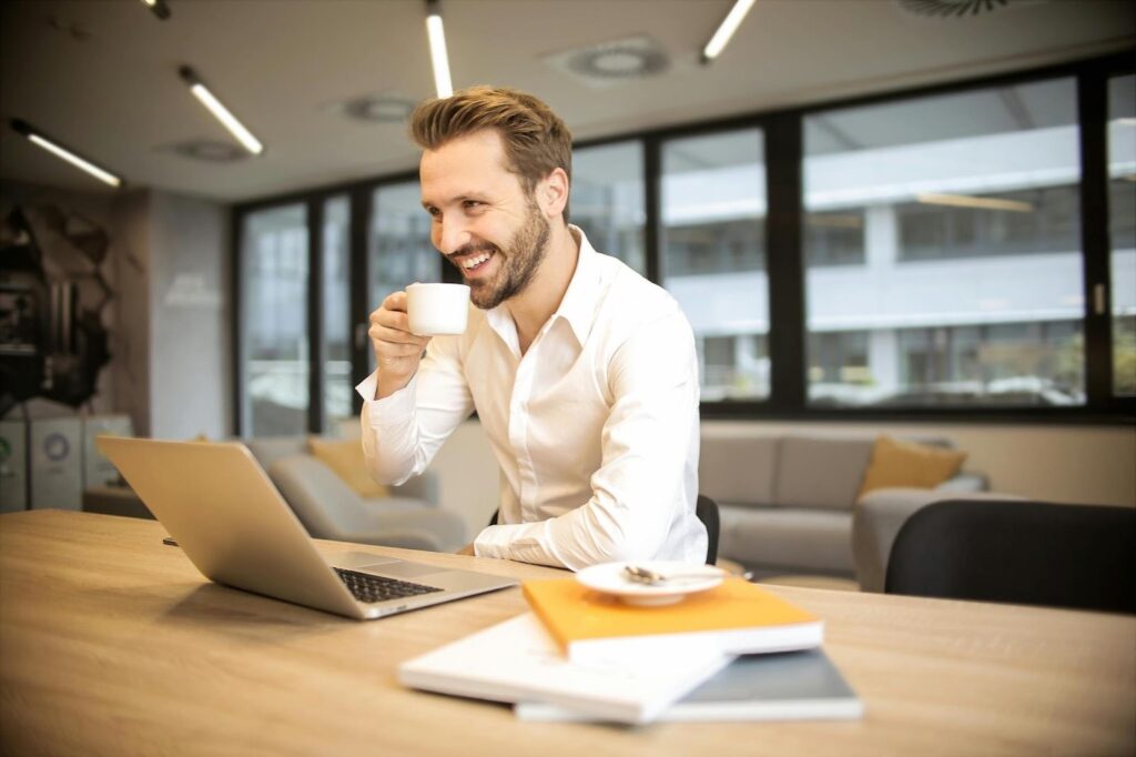 A smiling man in a white shirt sits at a desk with a laptop, holding a cup of coffee. There are papers, books, and a saucer on the desk, and large windows in the background let in natural light. | MONEY6X