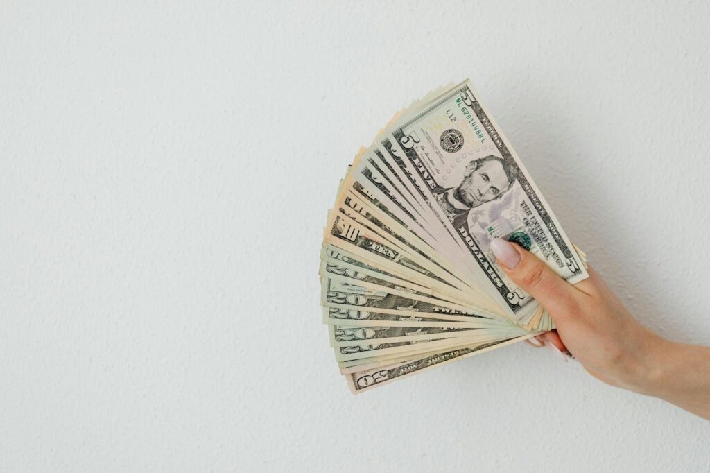 A hand holding a fanned-out stack of U.S. dollar bills against a plain white wall background. | MONEY6X