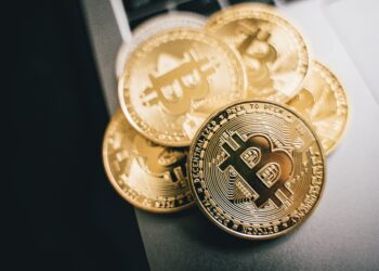 Several shiny gold Bitcoin coins with the Bitcoin symbol are stacked on a laptop keyboard, emphasizing the concept of digital currency and cryptocurrency technology. | MONEY6X