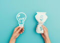 Two hands against a blue background: one holds a paper cut-out of a light bulb, symbolizing ideas, while the other holds a bag with a dollar sign, representing money or passive income. | MONEY6X