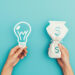 Two hands against a blue background: one holds a paper cut-out of a light bulb, symbolizing ideas, while the other holds a bag with a dollar sign, representing money or passive income. | MONEY6X