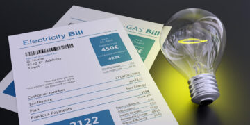 A close-up of electricity and gas bills next to a glowing light bulb on a dark surface, symbolizing energy usage and costs. The electricity bill shows the total amount due as 450 euros. | MONEY6X