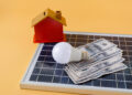 A small model house, a light bulb, and several U.S. hundred-dollar bills rest on a solar panel against a yellow background, symbolizing home energy savings with solar power. | MONEY6X