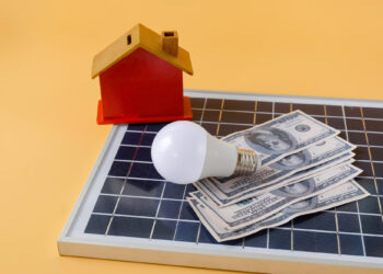 A small model house, a light bulb, and several U.S. hundred-dollar bills rest on a solar panel against a yellow background, symbolizing home energy savings with solar power. | MONEY6X