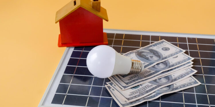 A small model house, a light bulb, and several U.S. hundred-dollar bills rest on a solar panel against a yellow background, symbolizing home energy savings with solar power. | MONEY6X