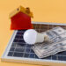 A small model house, a light bulb, and several U.S. hundred-dollar bills rest on a solar panel against a yellow background, symbolizing home energy savings with solar power. | MONEY6X