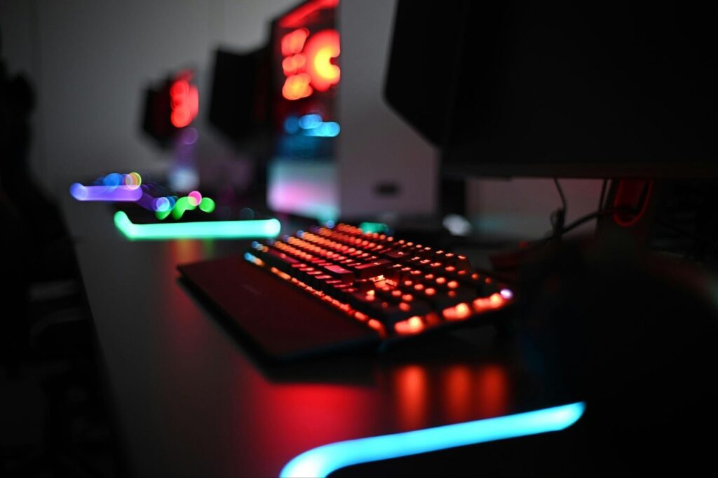 A close-up of a gaming keyboard with colorful RGB backlighting on a dark desk, next to other illuminated gaming setups, creating a vibrant and futuristic atmosphere. | MONEY6X