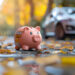 A pink piggy bank sits on a wet, leaf-covered street with a blurred silver car and autumn trees in the background, suggesting themes of savings and finances. | MONEY6X