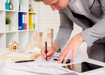 A person in a gray suit is drawing architectural plans on a desk with various tools, papers, small wooden house models, and a tablet, suggesting work related to architecture or real estate. | MONEY6X