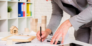 A person in a gray suit is drawing architectural plans on a desk with various tools, papers, small wooden house models, and a tablet, suggesting work related to architecture or real estate. | MONEY6X