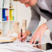 A person in a gray suit is drawing architectural plans on a desk with various tools, papers, small wooden house models, and a tablet, suggesting work related to architecture or real estate. | MONEY6X