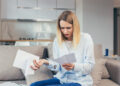 A woman sitting on a couch looks worried as she reads a paper bill, holding more documents in her other hand. She is in a modern living room with a kitchen in the background. | MONEY6X
