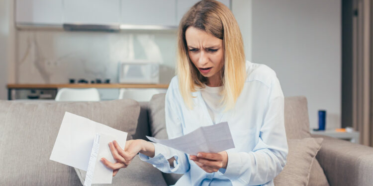 A woman sitting on a couch looks worried as she reads a paper bill, holding more documents in her other hand. She is in a modern living room with a kitchen in the background. | MONEY6X