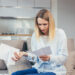 A woman sitting on a couch looks worried as she reads a paper bill, holding more documents in her other hand. She is in a modern living room with a kitchen in the background. | MONEY6X