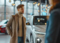 A man in a tan coat and turtleneck stands indoors near a white car, looking at another person. The setting appears to be a modern car showroom with large windows and hanging lights. | MONEY6X