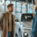 A man in a tan coat and turtleneck stands indoors near a white car, looking at another person. The setting appears to be a modern car showroom with large windows and hanging lights. | MONEY6X