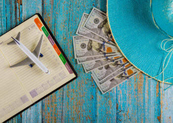 A model airplane on an open planner, several $100 bills, and a turquoise sunhat on a distressed blue wooden surface, suggesting travel planning and expenses. | MONEY6X
