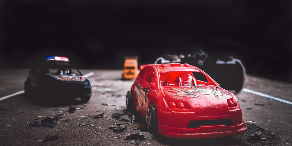 A close-up of a small red toy car appears damaged on a road, surrounded by toy police and other cars, with debris scattered on the dark surface. | MONEY6X