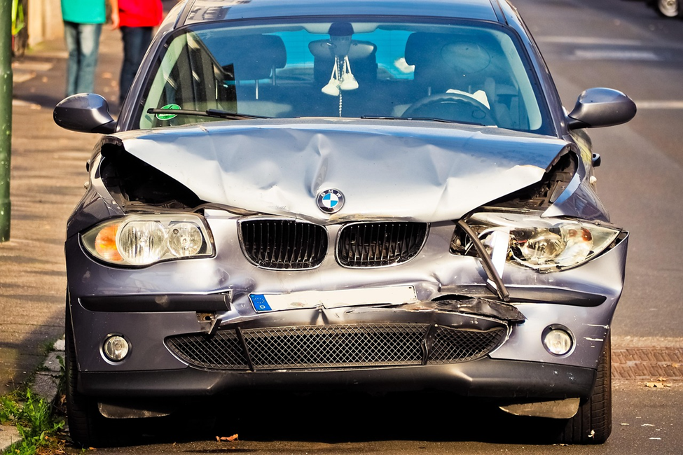 A gray BMW car with severe front-end damage is parked on the side of the street, indicating it was involved in a collision. The hood is crumpled and both headlights are broken. | MONEY6X
